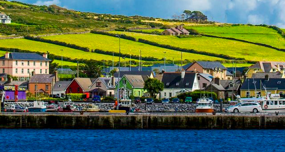 Colorful houses line a waterfront with boats moored in front; green hills and fields are visible in the background under a partly cloudy sky—capturing the charming scenery often experienced on Ireland tours.