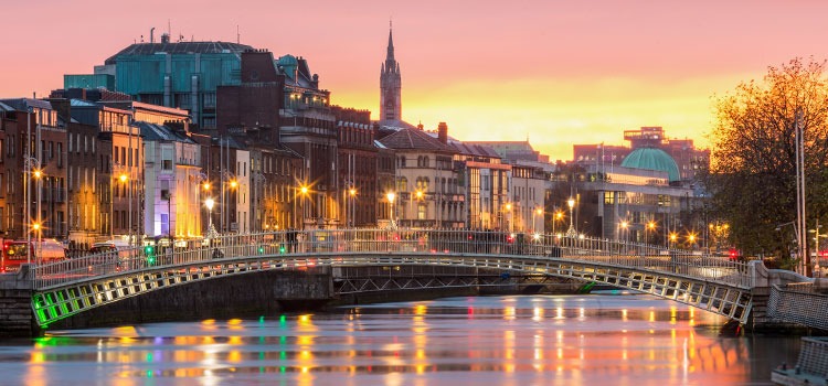 Ha’penny Bridge spans the River Liffey in Dublin at sunset, with historic buildings and city lights reflected in the water—a picture-perfect moment often enjoyed on Ireland tours or during 5 day tours exploring the vibrant heart of the city.