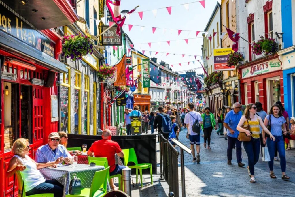 A busy pedestrian street with colorful shopfronts, outdoor café seating, hanging flower baskets, and people walking or dining on a sunny day. Pink bunting is strung above the street, creating a lively atmosphere often enjoyed by travelers on Ireland tours.