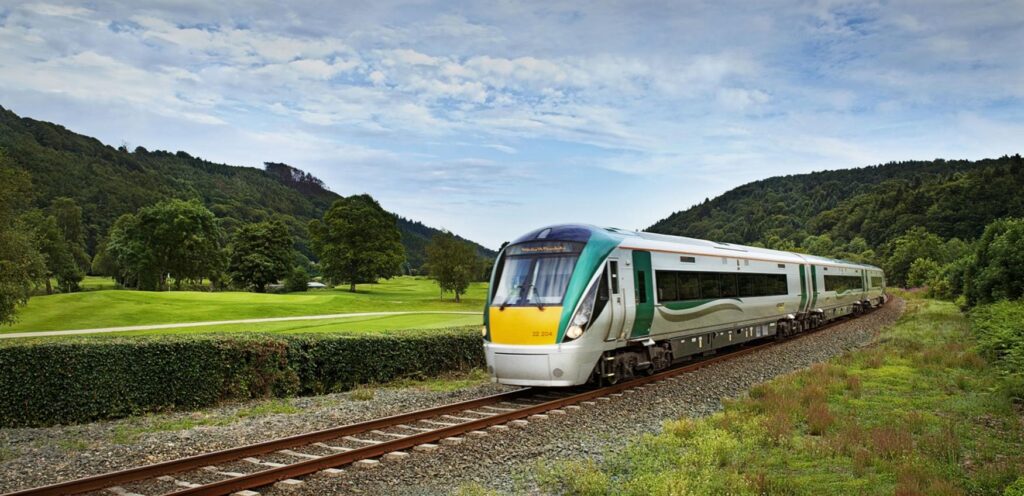 A modern passenger train travels along railway tracks through a green landscape with hills and trees under a partly cloudy sky, offering scenic views often featured on Ireland tours, including popular 10 day tours of Ireland.