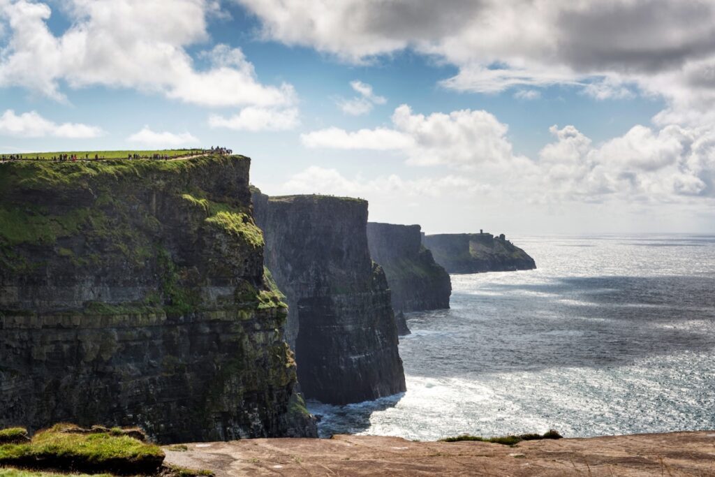 Steep sea cliffs rise above the Atlantic Ocean under a partly cloudy sky, with green grass on top and waves crashing below—the iconic Cliffs of Moher are a breathtaking highlight often included in a Connemara or Galway day tour.