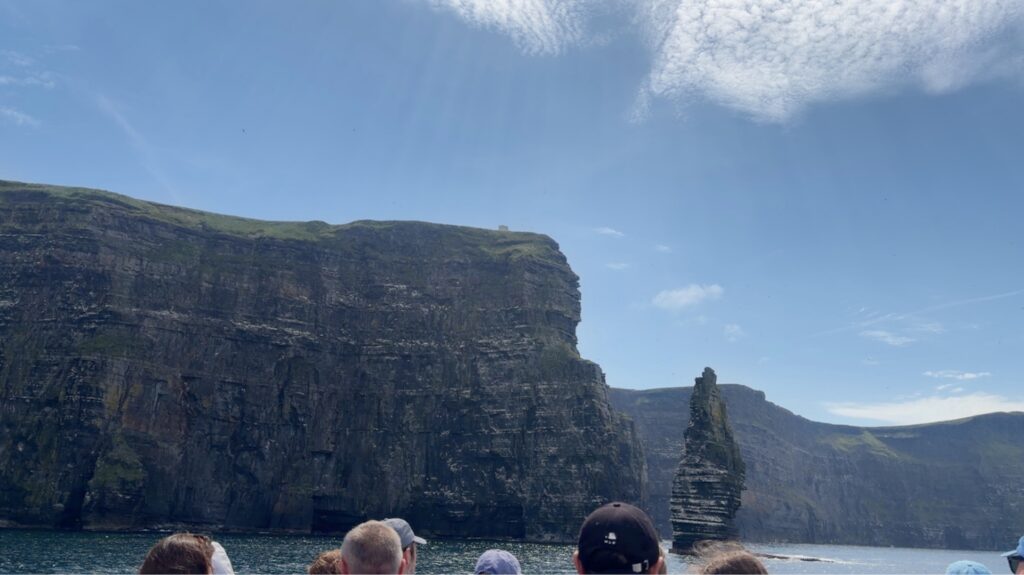 People on a boat look toward the Cliffs of Moher and a sea stack rising from the ocean under a partly cloudy sky, capturing the breathtaking scenery often enjoyed during a memorable Galway day tour.