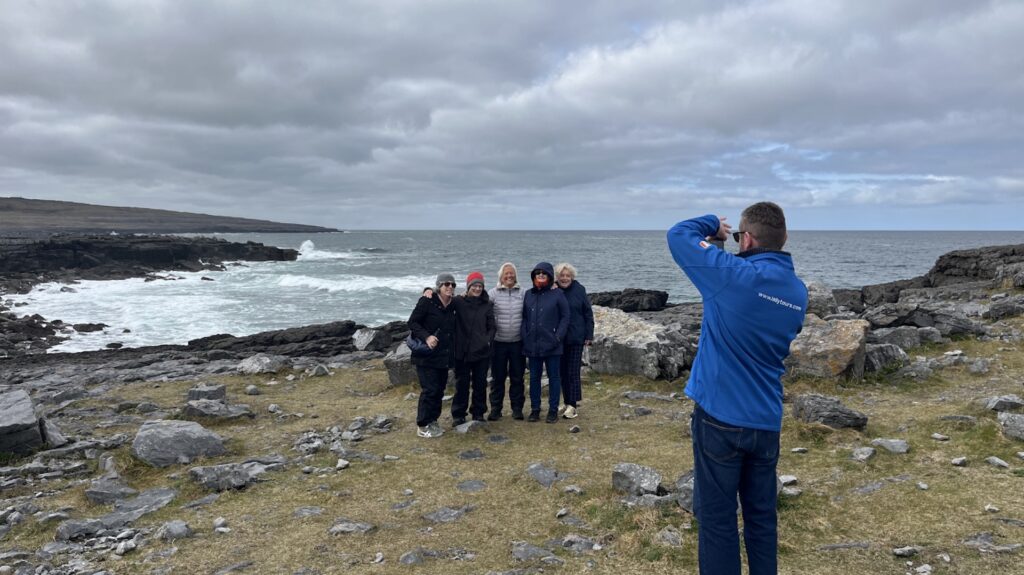 A person in a blue jacket takes a photo of five people standing together on a rocky coastline with the ocean and cloudy sky in the background, capturing memories during their Galway day tour.