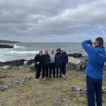 A person in a blue jacket takes a photo of five people standing together on a rocky coastline with the ocean and cloudy sky in the background, capturing memories during their Galway day tour.