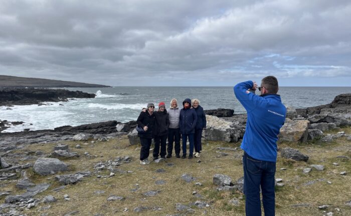 A person in a blue jacket takes a photo of five people standing together on a rocky coastline with the ocean and cloudy sky in the background, capturing memories during their Galway day tour.