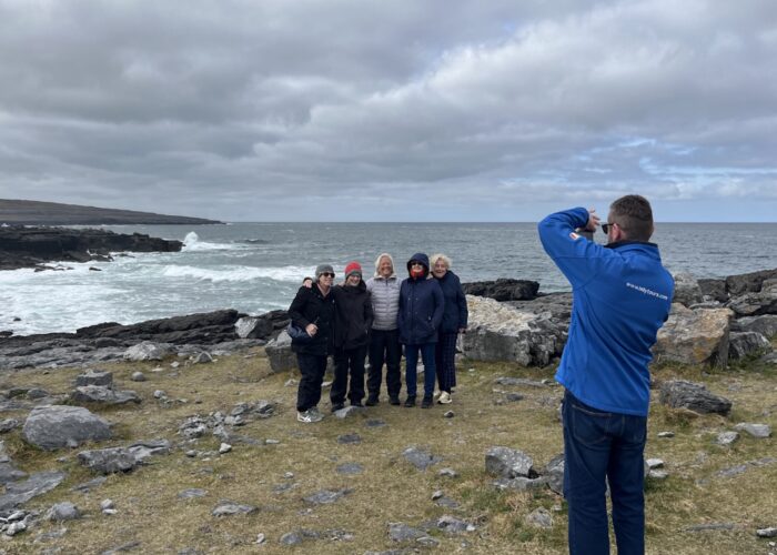 A person in a blue jacket takes a photo of five people standing together on a rocky coastline with the ocean and cloudy sky in the background, capturing memories during their Galway day tour.