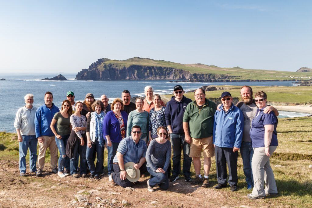A group of people poses for a photo on a grassy area by the sea, with cliffs and coastline in the background under a clear sky—capturing the scenic beauty often experienced on Ireland tours.