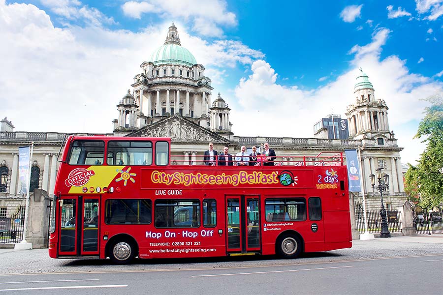 Red double-decker City Sightseeing bus parked in front of Belfast City Hall on a sunny day, with people seated on the open top deck—a perfect scene for those enjoying Ireland Tours or as part of an 8 Day Ireland Tour.