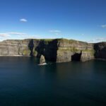 Cliffs of Moher rise above the Atlantic Ocean under a blue sky with scattered clouds; sharp rock formations jut from the water below—an iconic highlight on many Ireland tours.