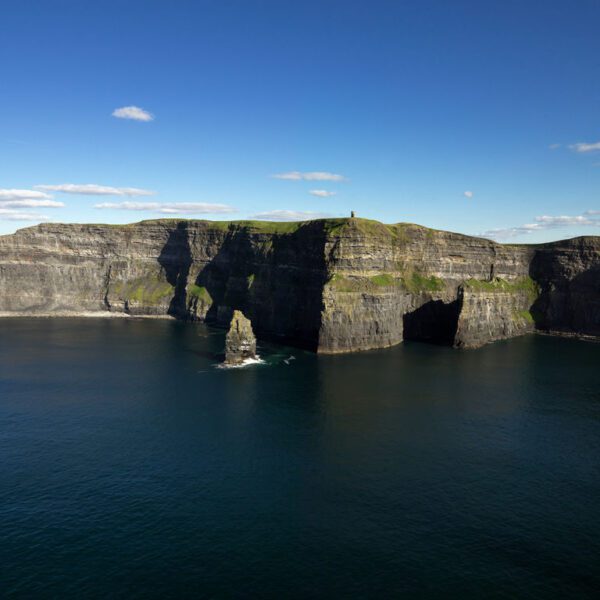 Cliffs of Moher rise above the Atlantic Ocean under a blue sky with scattered clouds; sharp rock formations jut from the water below—an iconic highlight on many Ireland tours.