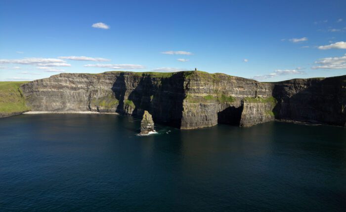 Cliffs of Moher rise above the Atlantic Ocean under a blue sky with scattered clouds; sharp rock formations jut from the water below—an iconic highlight on many Ireland tours.