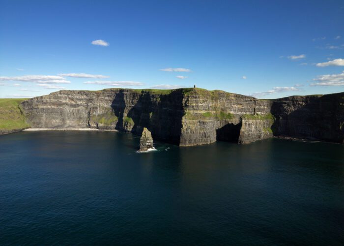 Cliffs of Moher rise above the Atlantic Ocean under a blue sky with scattered clouds; sharp rock formations jut from the water below—an iconic highlight on many Ireland tours.