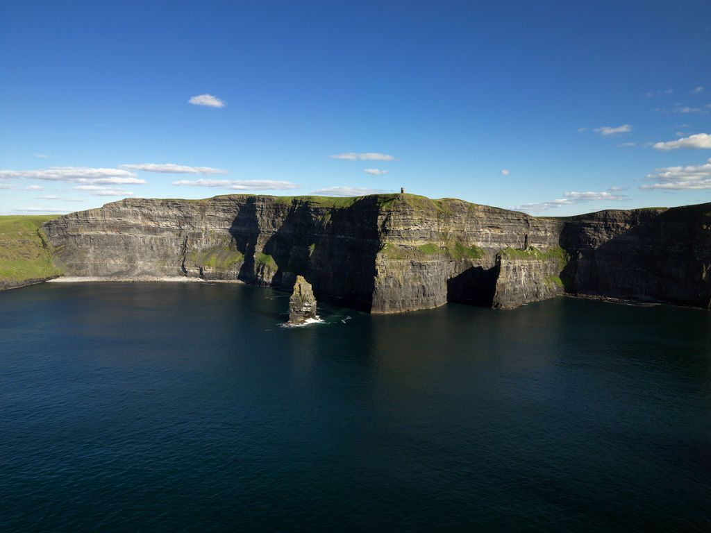 Cliffs of Moher rise above the Atlantic Ocean under a blue sky with scattered clouds; sharp rock formations jut from the water below—an iconic highlight on many Ireland tours.