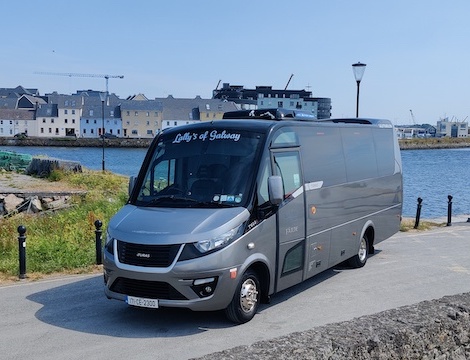 A grey tour bus labeled “Lally’s of Galway” is parked by a waterfront, with buildings and construction cranes visible in the background under a clear sky.