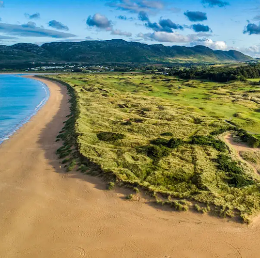 Aerial view of a sandy beach meeting grassy dunes, with rolling hills and scattered clouds in the background under a blue sky.