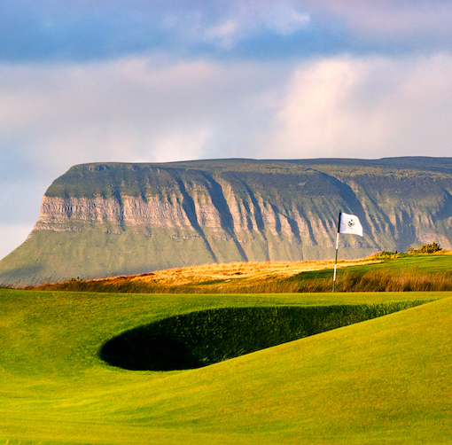 A golf green with a flag and sand bunker in the foreground, with a large, flat-topped mountain in the background under a partly cloudy sky.