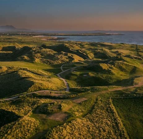 Aerial view of green hills with winding paths near a coastline under a clear sky at sunset.