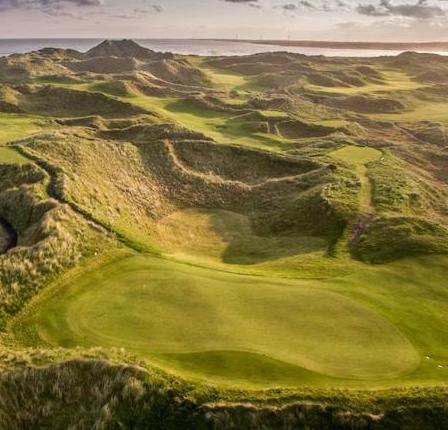 A golf course with rolling green fairways and sand dunes under a partly cloudy sky, with the sea visible in the distance.