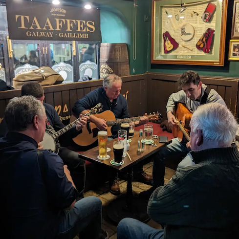 Five men sit around a small table playing guitars and a banjo in a cozy pub with drinks and musical decor on the walls.