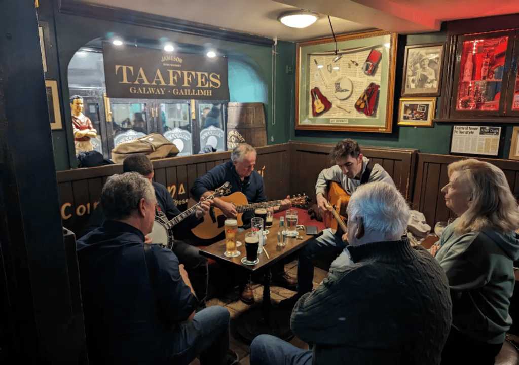 Five people sit around a table with drinks at a pub, three of them playing guitars. The pub has framed memorabilia and a sign reading “TAAFFES GALWAY – GAILLIMH.” It’s the perfect spot for travelers winding down after a day on Irish golf tours, capturing the spirit of Galway between rounds.