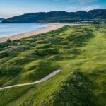 Aerial view of Northwest Links Golf, a stunning coastal golf course with rolling green fairways, sandy dunes, a beach, and hills in the background under a clear sky—an unforgettable highlight of the Golf Series by Lally Tours.
