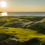 An Irish Links Golf course with rolling greens and sand traps sits beside the ocean at sunset, sunlight casting long shadows over the breathtaking landscape.