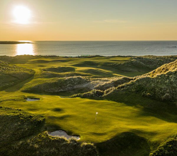 An Irish Links Golf course with rolling greens and sand traps sits beside the ocean at sunset, sunlight casting long shadows over the breathtaking landscape.
