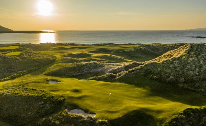 An Irish Links Golf course with rolling greens and sand traps sits beside the ocean at sunset, sunlight casting long shadows over the breathtaking landscape.