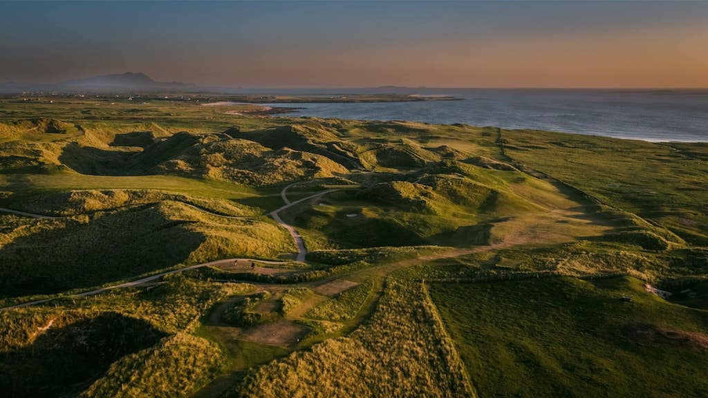 Aerial view of a coastal golf course in Ireland with rolling green hills, winding paths, and the ocean in the distance.