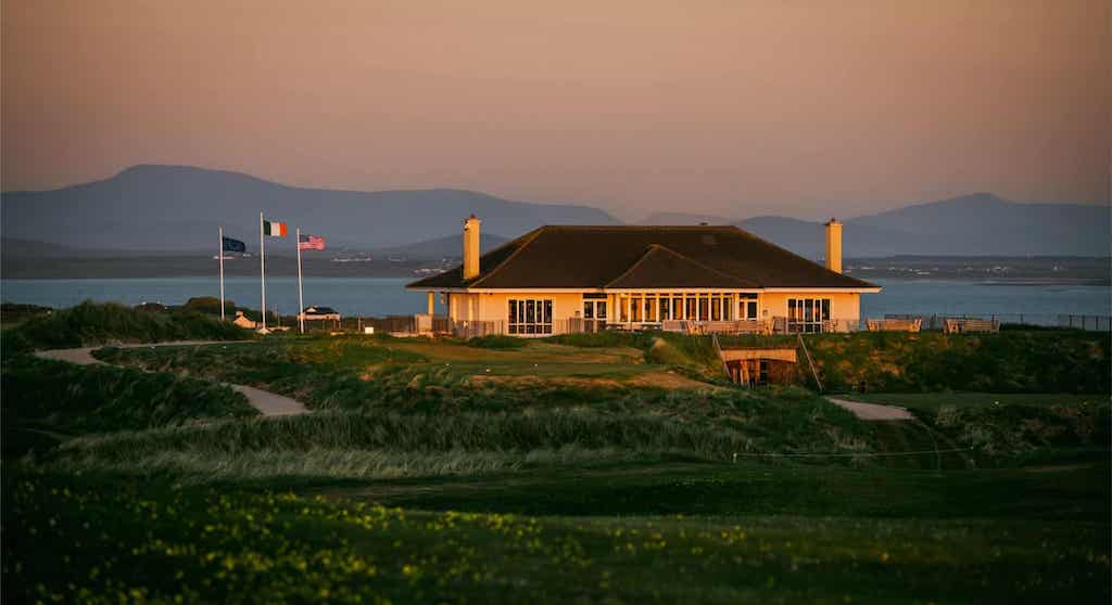 A clubhouse overlooking a golf course and bay at sunset with mountains in the distance, perfect for those planning to golf in Ireland.