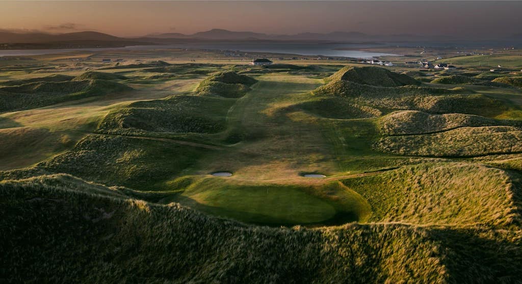An aerial view of a green golf course fairway nestled between grassy sand dunes, with a coastline and mountains in the distance, typical of golf in Ireland.