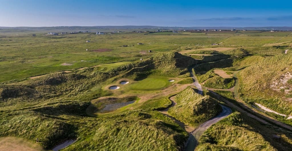An aerial view of a links golf course in Ireland, with the hole surrounded by sand dunes and rolling green hills.