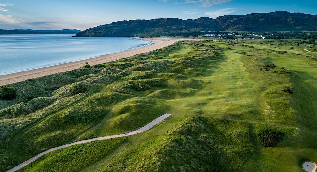Aerial view of a coastal golf course in Ireland with rolling green hills beside a sandy beach and ocean.
