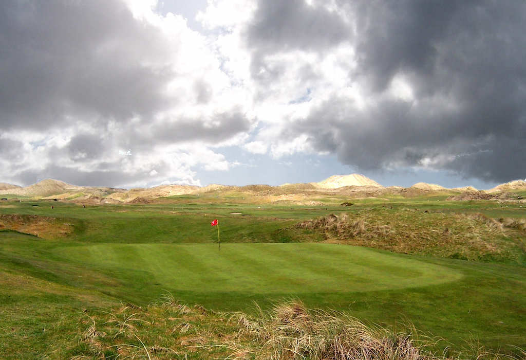 A golf green with a red flag set against grassy dunes under a cloudy sky, characteristic of golf in Ireland.