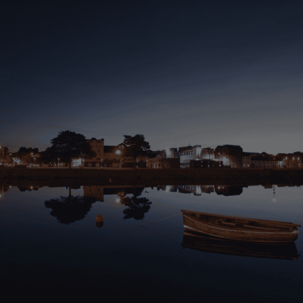 A wooden boat floats on calm water at twilight, reflecting the buildings and streetlights of Galway City, a popular destination for any walking tour.