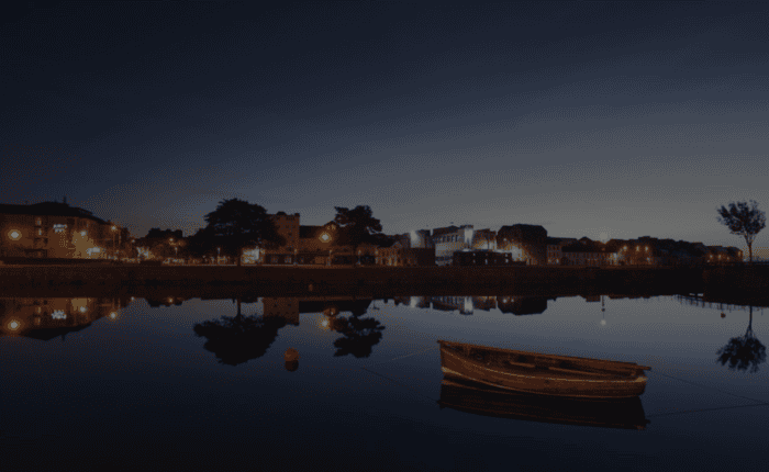 A wooden boat floats on calm water at twilight, reflecting the buildings and streetlights of Galway City, a popular destination for any walking tour.