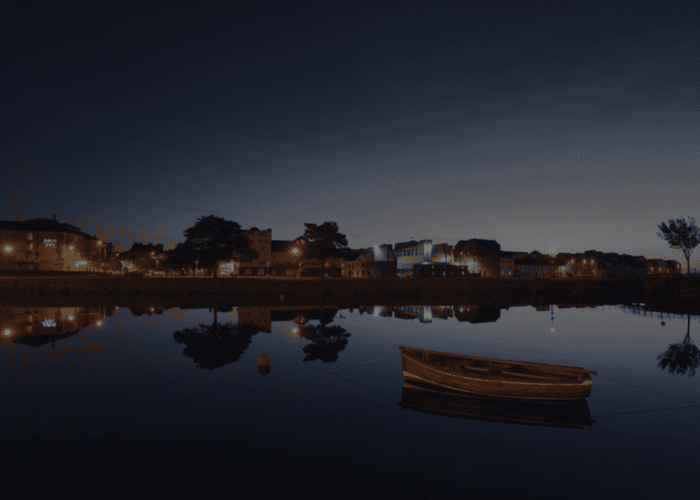 A wooden boat floats on calm water at twilight, reflecting the buildings and streetlights of Galway City, a popular destination for any walking tour.