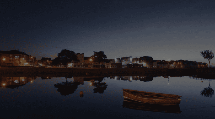 A wooden boat floats on calm water at twilight, reflecting the buildings and streetlights of Galway City, a popular destination for any walking tour.