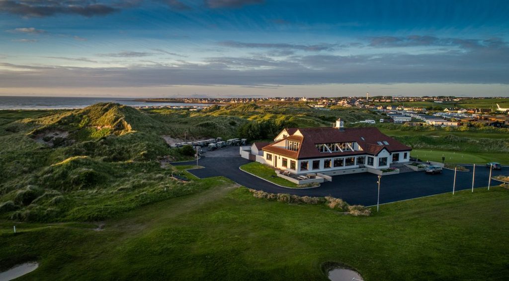 Aerial view of a golf clubhouse nestled among grassy dunes overlooking the coast and a town, an essential stop for an Ireland golf trip.