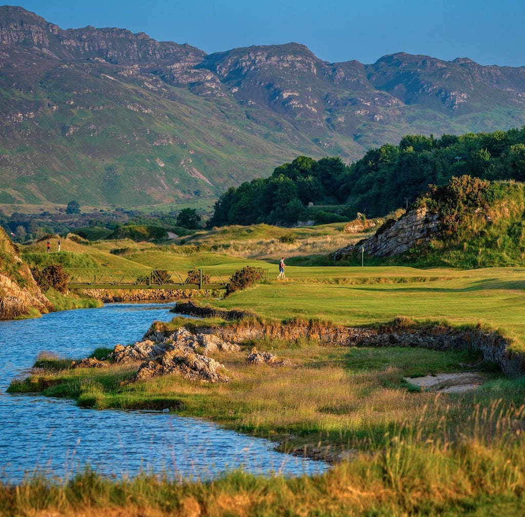 A golfer walks across a bridge on an Ireland golf trip, set amidst grassy hills and mountains.